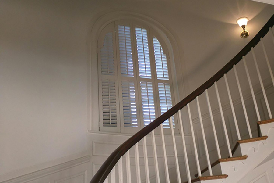Custom Polywood shutters fit every shape and size of stair windows. White Polywood shutters on an arched window in a stairwell